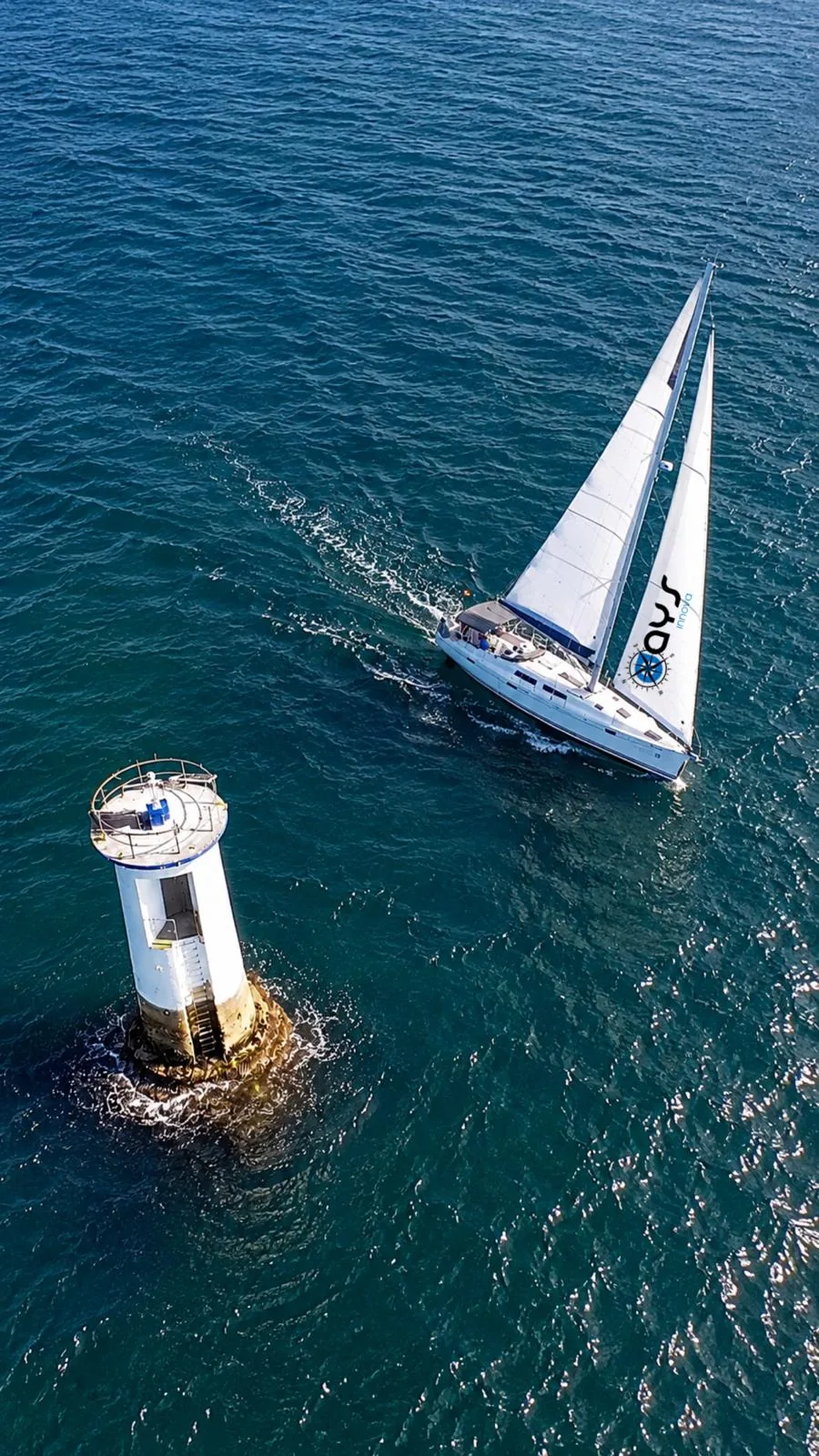 Velero con la vela de AYS Innova navegando junto a un faro en una ría gallega, vista aérea